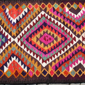 May include: A colorful rug with a geometric pattern in shades of red, orange, yellow, green, blue, purple, pink, and white. The rug has a diamond shape in the center and is made of woven fabric.