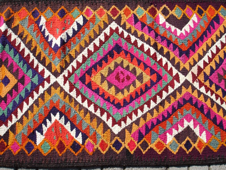 May include: A colorful rug with a geometric pattern in shades of red, orange, yellow, green, blue, purple, pink, and white. The rug has a diamond shape in the center and is made of woven fabric.