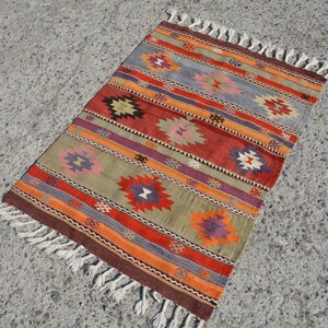 May include: A rectangular, woven rug with a geometric pattern. The rug features horizontal stripes in shades of red, orange, green, purple, and gray, with repeating diamond-like motifs. The edges are finished with a brown border and white fringe.