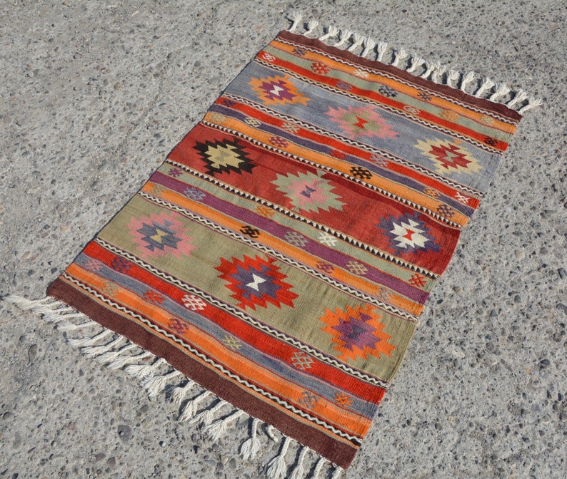 May include: A rectangular, woven rug with a geometric pattern. The rug features horizontal stripes in shades of red, orange, green, purple, and gray, with repeating diamond-like motifs. The edges are finished with a brown border and white fringe.