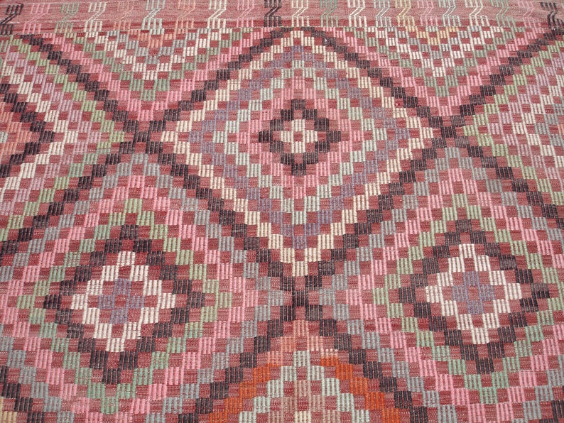 K&ouml;nnte beinhalten: Nahaufnahme eines rosafarbenen, gr&uuml;nen und schwarzen Teppichs mit geometrischem Muster. Der Teppich hat eine gewebte Textur und eine Diamantform in der Mitte.