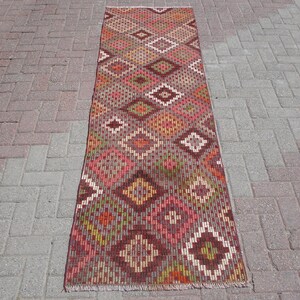 May include: A long, narrow rug with a geometric pattern in shades of red, brown, green, and white. The rug has a fringe border.