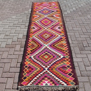 May include: A long, colorful rug with a geometric pattern in shades of red, pink, orange, yellow, green, blue, and brown. The rug has a fringe on both ends.