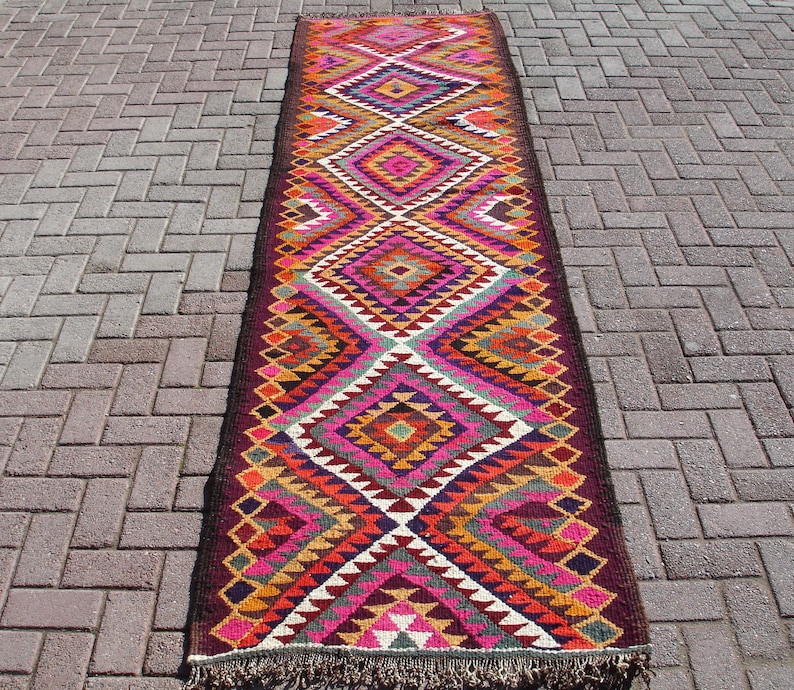 May include: A long, colorful rug with a geometric pattern in shades of red, pink, orange, yellow, green, blue, and brown. The rug has a fringe on both ends.