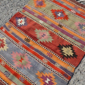 May include: A rectangular, hand-woven rug with a striped pattern. The rug features alternating bands of color, including red, orange, purple, and blue. Geometric floral motifs are interspersed throughout the design. The rug has a fringed edge.