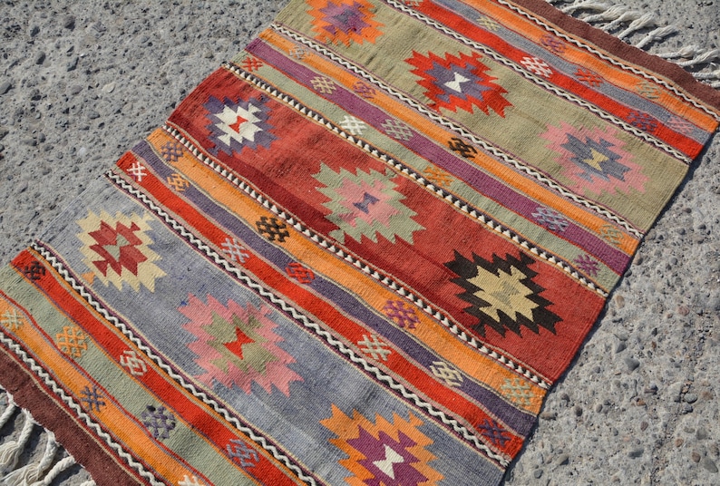 May include: A rectangular, hand-woven rug with a striped pattern. The rug features alternating bands of color, including red, orange, purple, and blue. Geometric floral motifs are interspersed throughout the design. The rug has a fringed edge.