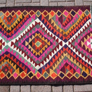 May include: A colorful rug with a geometric pattern in shades of pink, red, orange, yellow, green, blue, purple, and brown. The rug has a fringe border.