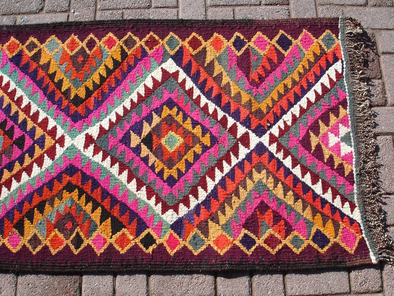 May include: A colorful rug with a geometric pattern in shades of pink, red, orange, yellow, green, blue, purple, and brown. The rug has a fringe border.