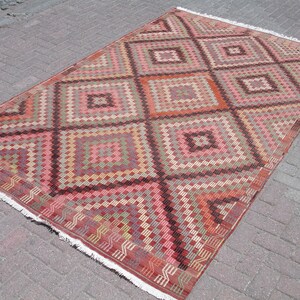 K&ouml;nnte beinhalten: Ein antiker t&uuml;rkischer Teppich mit einem geometrischen Muster in Rot-, Rosa-, Braun- und Cremet&ouml;nen. Der Teppich hat einen Fransenrand und ist in traditioneller Kilim-Technik gewebt.