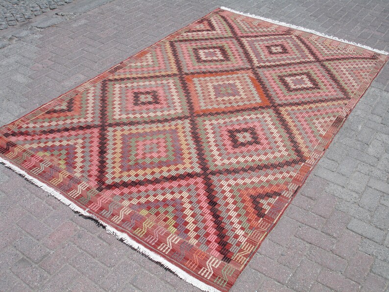 K&ouml;nnte beinhalten: Ein antiker t&uuml;rkischer Teppich mit einem geometrischen Muster in Rot-, Rosa-, Braun- und Cremet&ouml;nen. Der Teppich hat einen Fransenrand und ist in traditioneller Kilim-Technik gewebt.