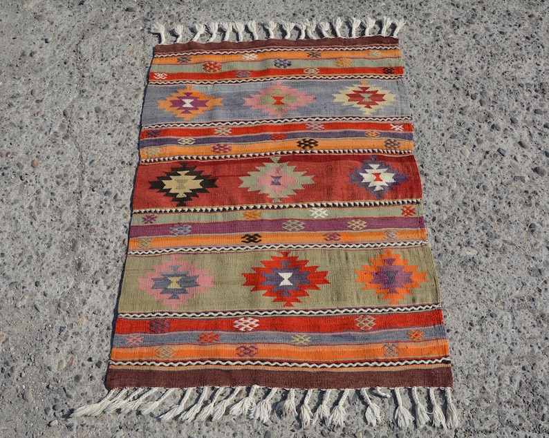 May include: A rectangular, hand-woven rug with a geometric pattern. The rug features a brown border and fringe, with alternating stripes of orange, red, green, and purple. The design includes diamond-shaped motifs in various colors.