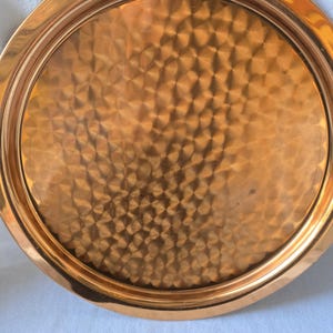 Może przedstawiać: Okrągła miedziana taca do serwowania z młotkowanym wykończeniem. Taca ma podniesiony rant i gładką, polerowaną powierzchnię.