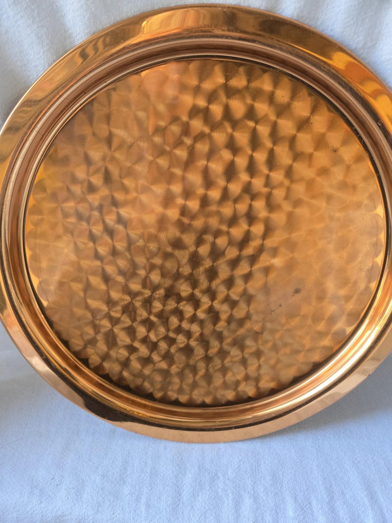 Może przedstawiać: Okrągła miedziana taca do serwowania z młotkowanym wykończeniem. Taca ma podniesiony rant i gładką, polerowaną powierzchnię.