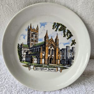 May include: A white ceramic plate featuring a detailed illustration of Buckfast Abbey. The image showcases the abbey's architecture with brown, orange, and green hues, set against a blue sky. The plate has a simple, rounded edge.