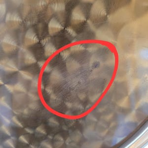 Może przedstawiać: Zbliżenie na stalowy garnek ze śladem po zarysowaniu w kształcie koła na dnie.