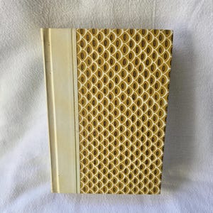 May include: A hardcover book with a cream-colored spine and a patterned cover. The cover features a repeating design of brown ovals on a gold background. The book's spine is a solid cream color.