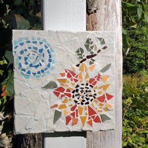 May include: A mosaic tile art piece featuring a stylized flower with red, orange, and yellow petals. The flower is surrounded by a blue and white swirl design.