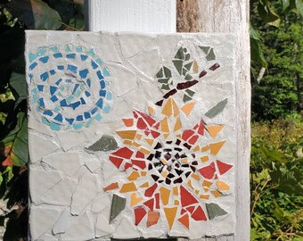 May include: A mosaic tile art piece featuring a stylized flower with red, orange, and yellow petals. The flower is surrounded by a blue and white swirl design.