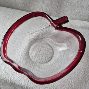 May include: A clear glass apple-shaped bowl with a deep red rim and stem. The bowl has a delicate leaf design etched into the glass. The bowl is likely used for serving or display.