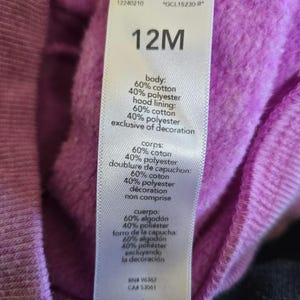 Może przedstawiać: Fioletowa bluza z kapturem z białą metką, na kt&oacute;rej widnieje napis "12M" i podany jest skład materiału: 60% bawełna i 40% poliester. Metka zawiera r&oacute;wnież instrukcje dotyczące pielęgnacji.