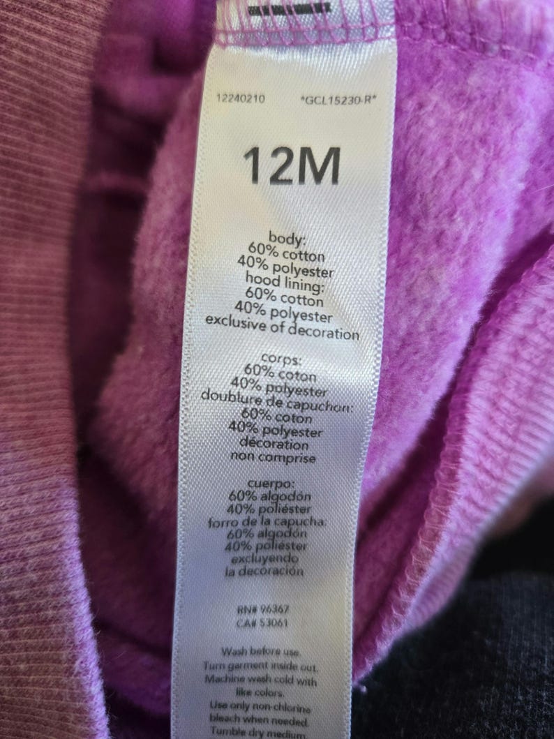 Może przedstawiać: Fioletowa bluza z kapturem z białą metką, na kt&oacute;rej widnieje napis "12M" i podany jest skład materiału: 60% bawełna i 40% poliester. Metka zawiera r&oacute;wnież instrukcje dotyczące pielęgnacji.