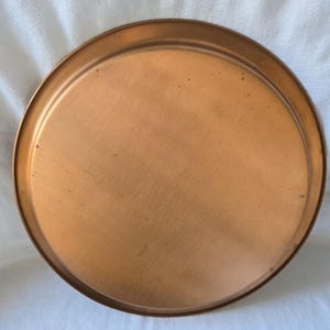 Może przedstawiać: Okrągła miedziana taca do serwowania z lekko uniesionym rantem. Taca jest pusta i ma błyszczące, metaliczne wykończenie.