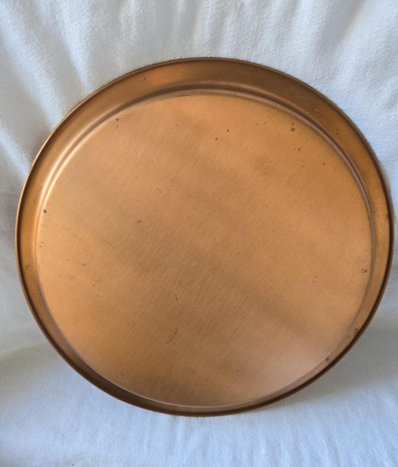 Może przedstawiać: Okrągła miedziana taca do serwowania z lekko uniesionym rantem. Taca jest pusta i ma błyszczące, metaliczne wykończenie.
