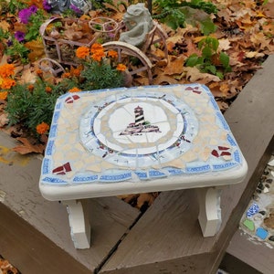 May include: A square mosaic tile table with a lighthouse design in the center. The table has a white painted frame and legs. The mosaic tiles are in shades of blue, beige, and red. The table is sitting on a brown wooden surface.