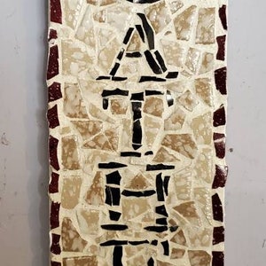 May include: A brown and white mosaic tile sign with the word "CATER" spelled out in black letters. The sign has a red border and a gold loop at the top.