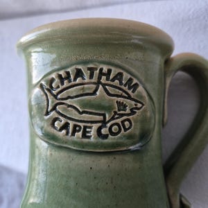 May include: Green ceramic mug with a handle. The mug features an embossed design with the words "CHATHAM CAPE COD" surrounding a shark illustration. The mug has a light brown rim and handle.