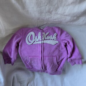Może przedstawiać: Fioletowa bluza z kapturem i zamkiem błyskawicznym z białym logo OshKosh z przodu.