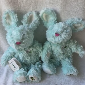 May include: Two plush stuffed bunnies with shaggy light blue fur, long ears with white inner linings, and small pink noses. One bunny features a white tag with the text "Gund" and a green ribbon graphic, while both have green ribbon accents.