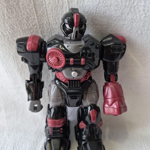 May include: A black and red toy robot with a detailed design. The robot has a helmet, chest plate, and articulated limbs. The robot's chest features a circular red design. The toy is made of plastic.
