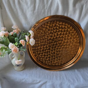 Może przedstawiać: Okrągła miedziana taca do serwowania z młotkowanym wykończeniem. Taca ma podniesiony rant i idealnie nadaje się do serwowania przekąsek, napoj&oacute;w lub innych przedmiot&oacute;w.