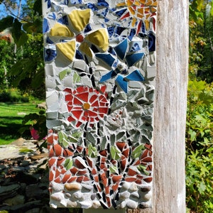 May include: A mosaic art piece depicting a cluster of flowers in shades of red, yellow, blue, and green. The flowers are made from broken pieces of ceramic tile and are set in a white mortar.