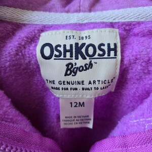Może przedstawiać: Fioletowa bluza z kapturem z białym zamkiem błyskawicznym i białą metką z logo OshKosh B'Gosh. Metka zawiera napis "EST. 1895" i "THE GENUINE ARTICLE MADE FOR FUN. BUILT TO LAST." Metka zawiera r&oacute;wnież napis "12M" i "MADE IN VIETNAM FABRIQUE AU VIETNAM HECHO EN VIETNAM."