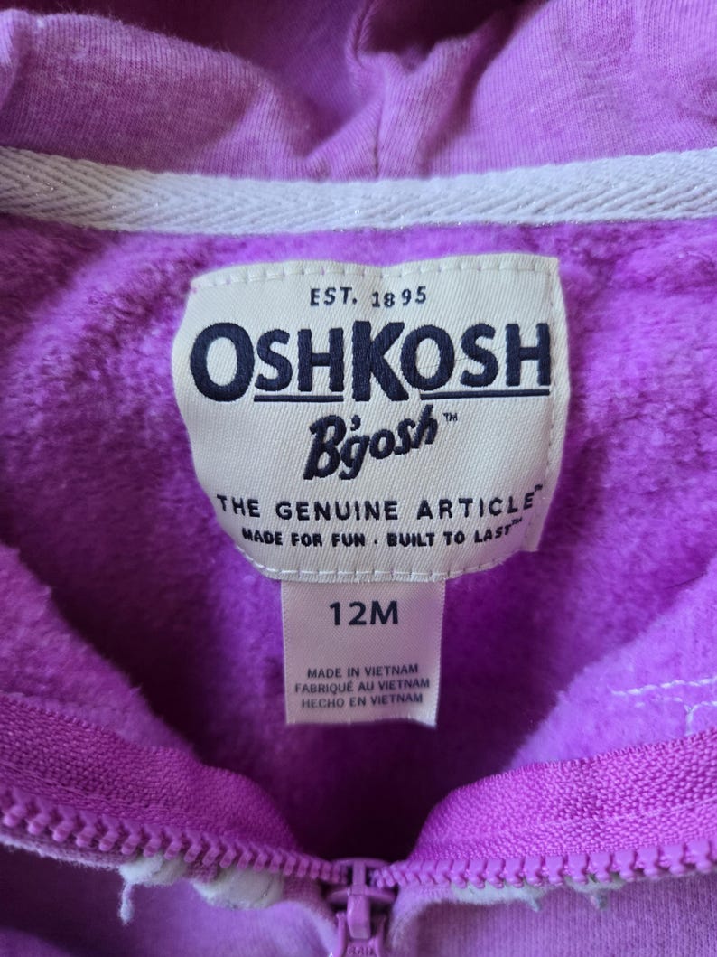 Może przedstawiać: Fioletowa bluza z kapturem z białym zamkiem błyskawicznym i białą metką z logo OshKosh B'Gosh. Metka zawiera napis "EST. 1895" i "THE GENUINE ARTICLE MADE FOR FUN. BUILT TO LAST." Metka zawiera r&oacute;wnież napis "12M" i "MADE IN VIETNAM FABRIQUE AU VIETNAM HECHO EN VIETNAM."