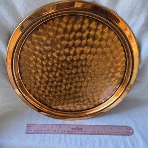 Może przedstawiać: Okrągła miedziana taca do serwowania z młotkowanym wykończeniem. Taca ma podniesiony rant i ma około 30 cm średnicy.