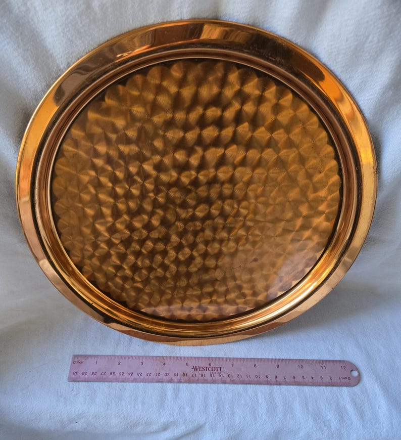 Może przedstawiać: Okrągła miedziana taca do serwowania z młotkowanym wykończeniem. Taca ma podniesiony rant i ma około 30 cm średnicy.