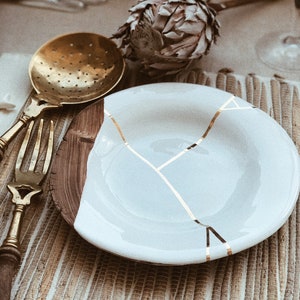 May include: A white ceramic plate with gold crackle lines, a brass strainer, and a fork on a woven placemat.