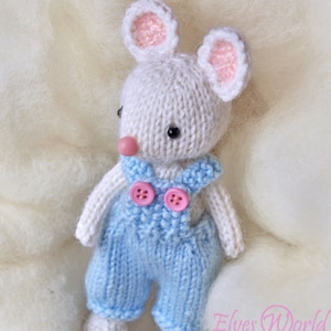 Könnte beinhalten: Eine weiße gestrickte Maus, die eine blaue Latzhose mit rosa Knöpfen trägt. Die Maus hat rosa Ohren und eine rosa Nase. Die Maus sitzt auf einer weißen, flauschigen Oberfläche. Das Bild enthält den Text "Elves World Toys & Patterns".