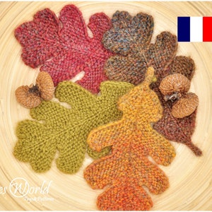 Peut inclure: Cinq feuilles de chêne tricotées de différentes couleurs, dont le rouge, le vert, le marron et l'orange, sont disposées sur une surface en bois. Les feuilles sont toutes de tailles et de formes différentes. Un petit drapeau français se trouve dans le coin supérieur droit de l'image.
