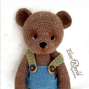 May include: A brown crocheted teddy bear wearing blue dungarees with yellow accents and brown buttons. The bear has black eyes and a black nose. The background is a white wall with a floral pattern.