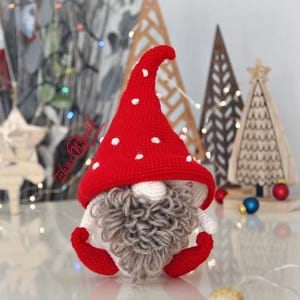 Puede incluir: Un gnomo hecho a mano con ganchillo con un gorro rojo con lunares blancos, una barba gris y pies rojos. El gnomo es una decoración festiva, perfecta para Navidad o decoración navideña. Otros árboles de Navidad de madera están en el fondo.