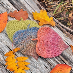 K&ouml;nnte beinhalten: Drei gestrickte Untersetzer in Blattform in Gr&uuml;n-, Blau- und Rosat&ouml;nen. Die Untersetzer sind auf einer Holzoberfl&auml;che mit Herbstbl&auml;ttern drumherum angeordnet.