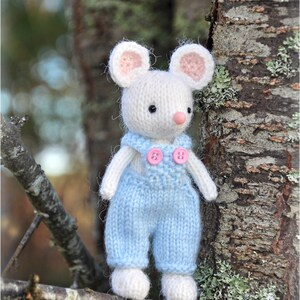 K&ouml;nnte beinhalten: Eine wei&szlig;e gestrickte Maus, die eine blaue Latzhose mit rosa Kn&ouml;pfen tr&auml;gt. Die Maus sitzt auf einem Ast mit einem Baumstamm im Hintergrund.