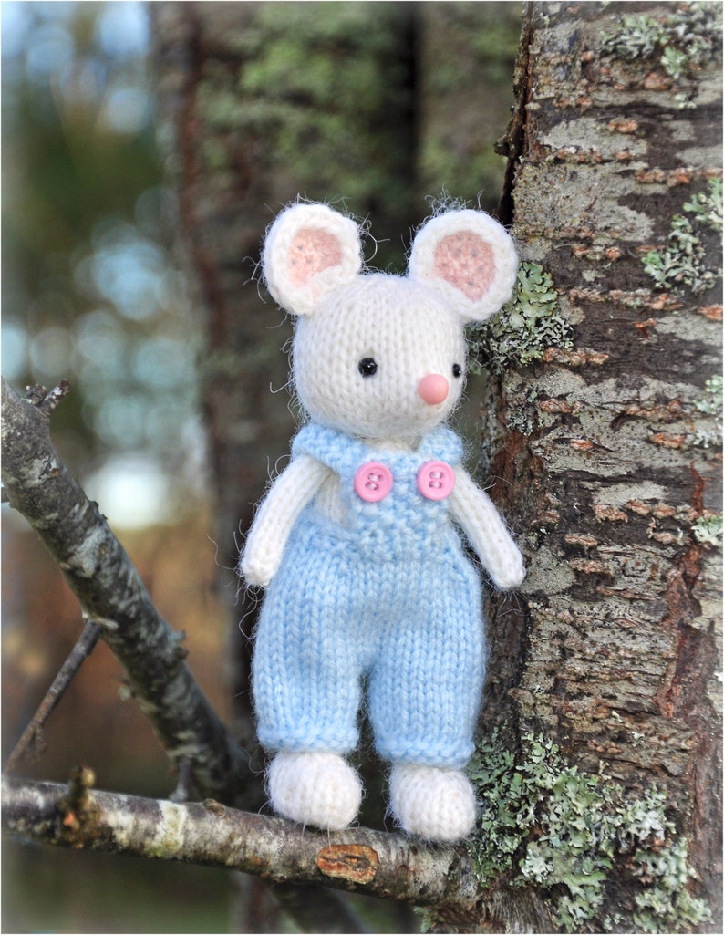 K&ouml;nnte beinhalten: Eine wei&szlig;e gestrickte Maus, die eine blaue Latzhose mit rosa Kn&ouml;pfen tr&auml;gt. Die Maus sitzt auf einem Ast mit einem Baumstamm im Hintergrund.