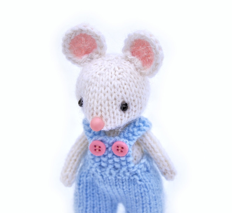 K&ouml;nnte beinhalten: Eine wei&szlig;e gestrickte Maus, die eine blaue Latzhose mit rosa Kn&ouml;pfen tr&auml;gt. Die Maus hat rosa Ohren und schwarze Augen.