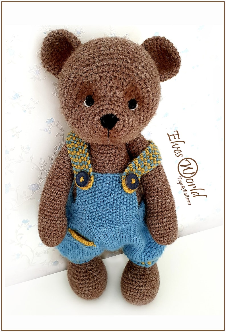 May include: A brown crocheted teddy bear wearing blue dungarees with yellow accents and brown buttons. The bear has black eyes and a black nose. The background is a white wall with a floral pattern.