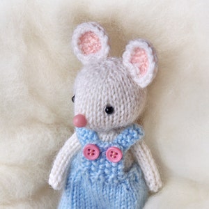 K&ouml;nnte beinhalten: Eine wei&szlig;e gestrickte Maus, die eine blaue Latzhose mit rosa Kn&ouml;pfen tr&auml;gt. Die Maus hat rosa Ohren und eine rosa Nase.